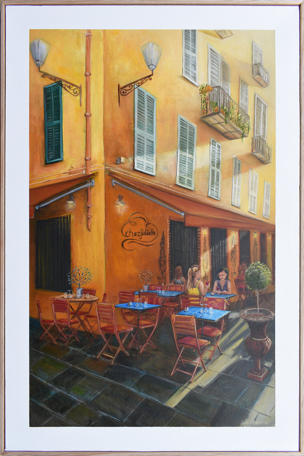 A painting of a yellow building with French window shutters and a cafe underneath the awnings of the streets of Nice, France hanging on a wall by Australian artist Jenni Rogers