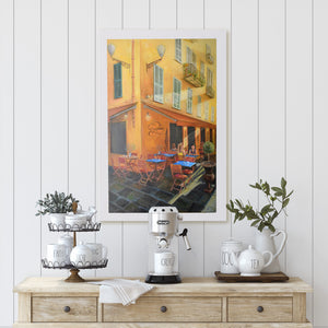 A painting of a yellow building with French window shutters and a cafe underneath the awnings of the streets of Nice, France hanging on a wall by Australian artist Jenni Rogers